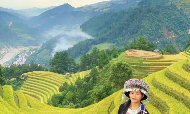 Hmong girl dressed in traditional attire standing amidst lush rice terraces with mountain backdrop.