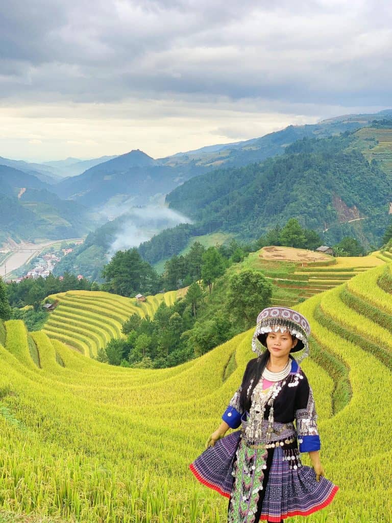 Hmong girl dressed in traditional attire standing amidst lush rice terraces with mountain backdrop.