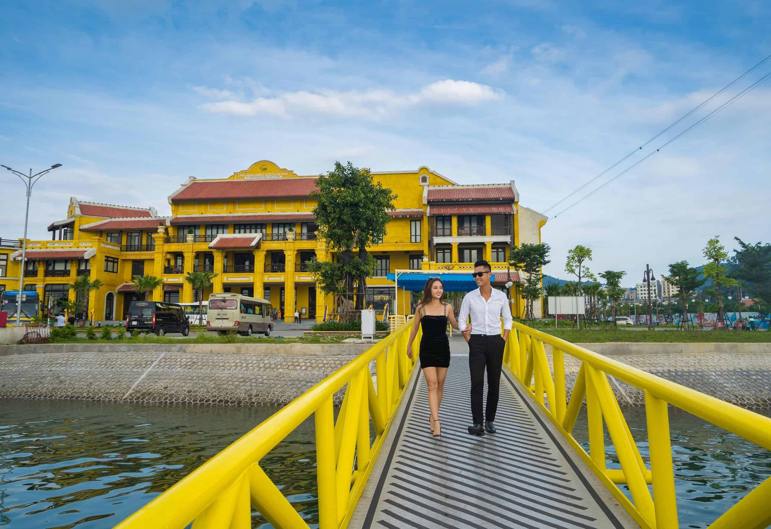 Romantic couple walking on a yellow bridge with Halong Bay scenery and cruise ship in background.