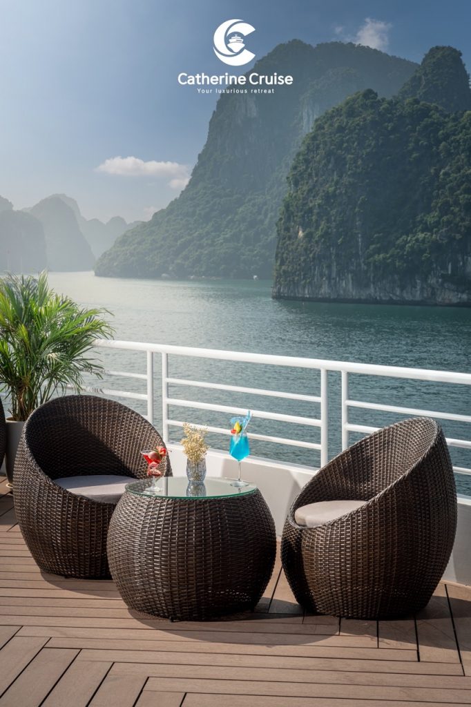 Luxury cruise deck with scenic bay view, wicker chairs, tropical drinks, and mountain landscape.
