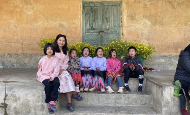 Children and local woman sitting on stone steps in a traditional Vietnamese village, vibrant colors.