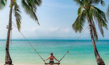 Beach scene with a woman on a swing between palm trees, turquoise sea, and clear sky, perfect for re.