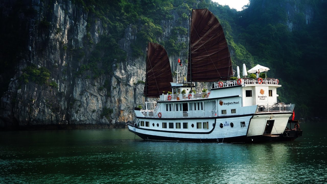 Scenic Bai Tu Long Bay cruise ship with traditional sails navigating lush green cliffs.