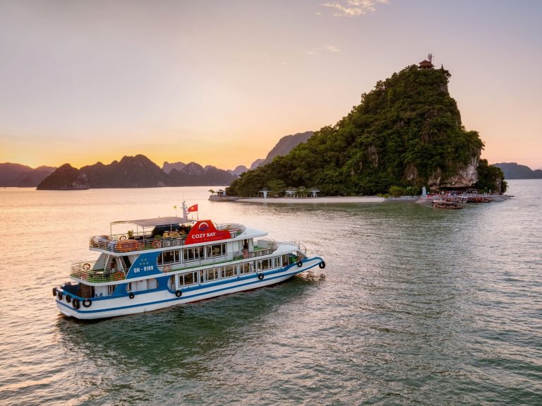 Beautiful scene of a luxury cruise boat sailing past lush limestone islands at sunset in Ha Long Bay.