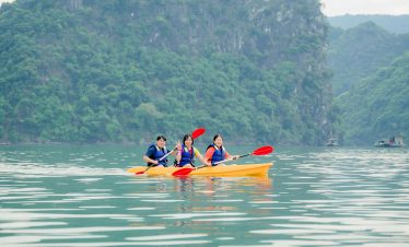 Kayaking scene on Halong Bay featuring three travelers paddling on calm waters with lush green cliff.