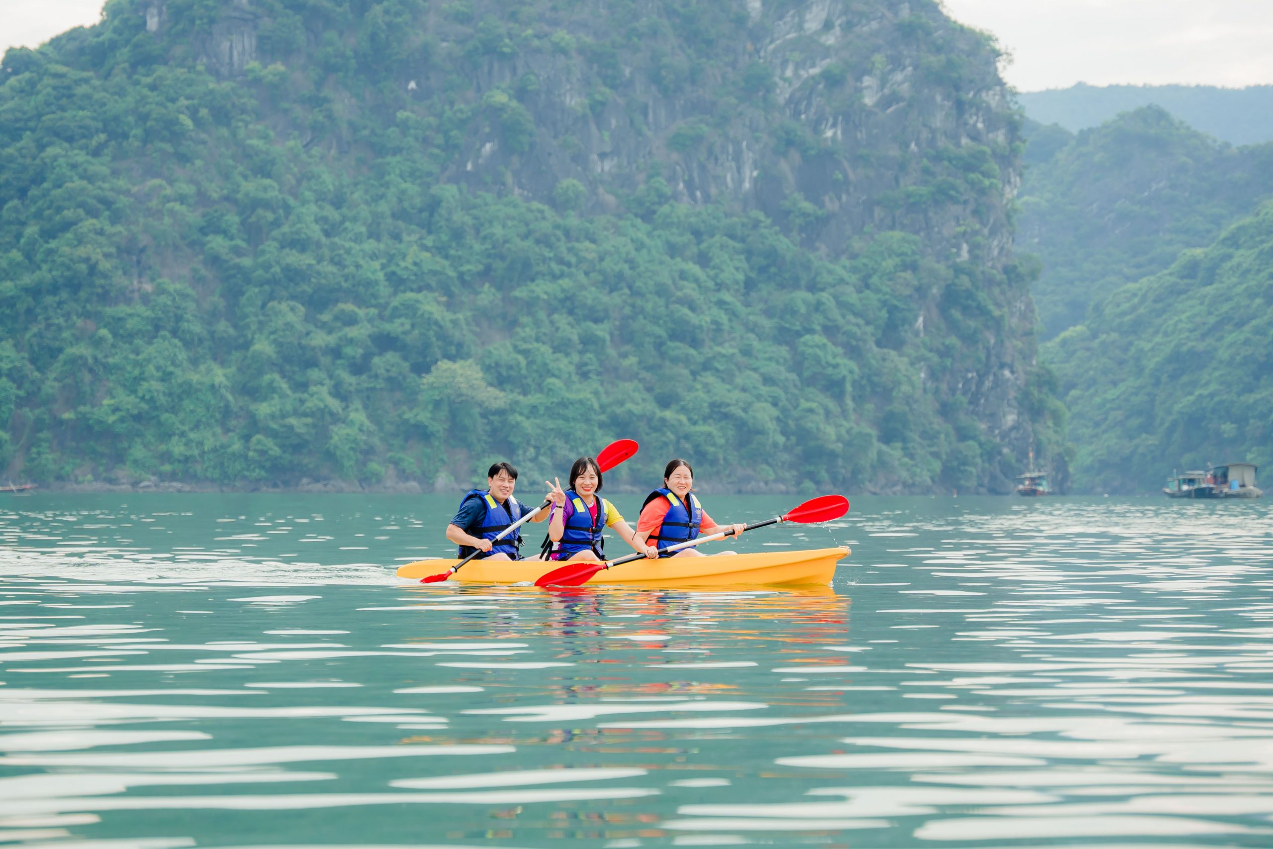 Kayaking scene on Halong Bay featuring three travelers paddling on calm waters with lush green cliff.