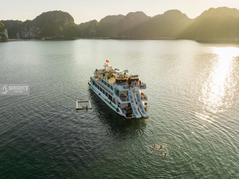 A cruise boat sailing on Halong Bay's calm waters during sunset, surrounded by towering limestone fo.
