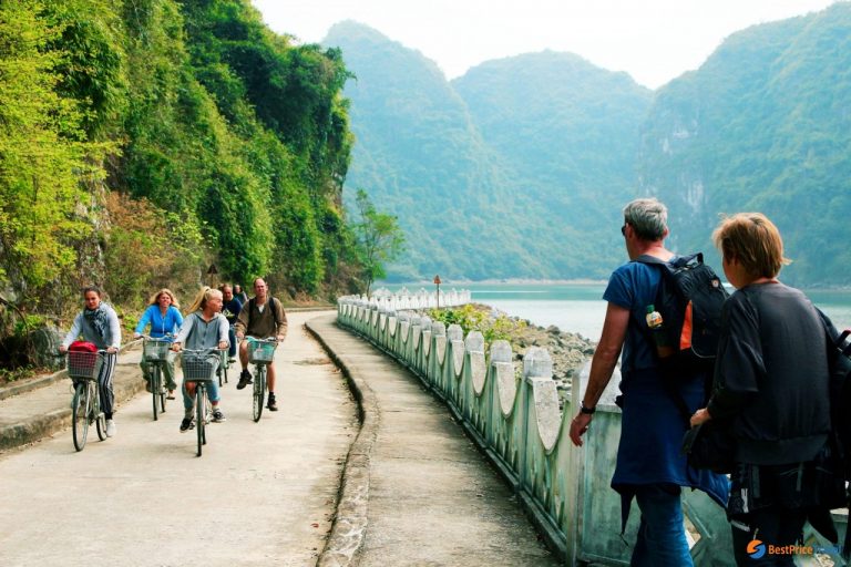 Cyclists enjoying scenic ride along river in Hanoi, Vietnam, with lush green mountains in background.