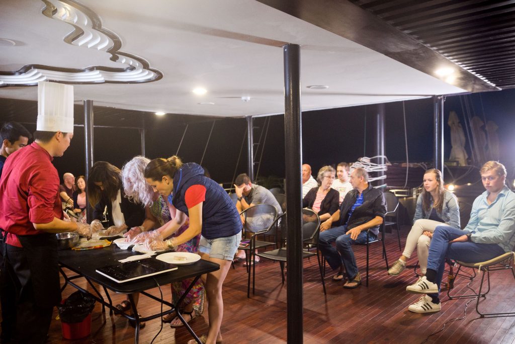 Cooking class on a boat with chefs and tourists enjoying a Vietnamese culinary experience at night.