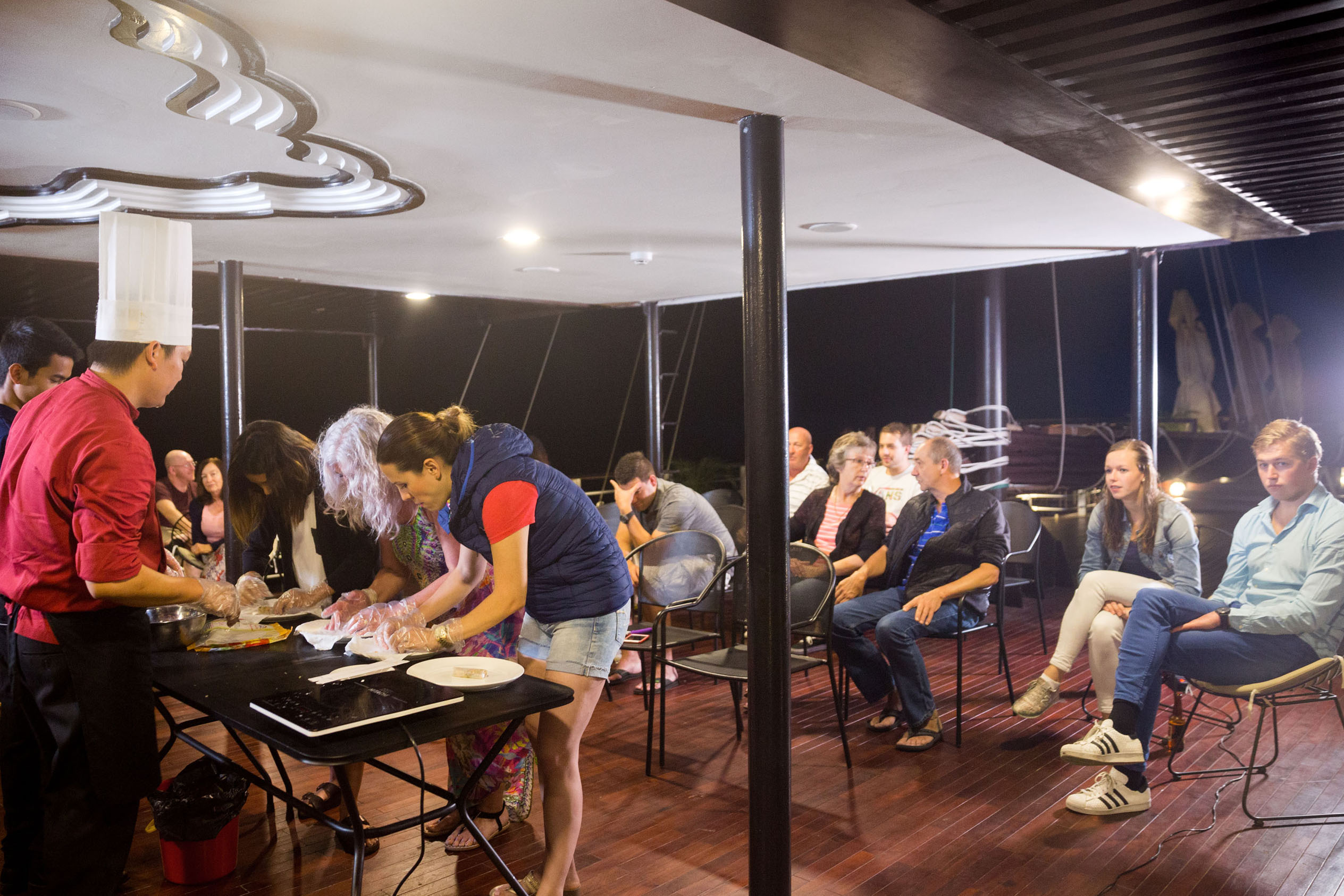 Cooking class on a boat with chefs and tourists enjoying a Vietnamese culinary experience at night.