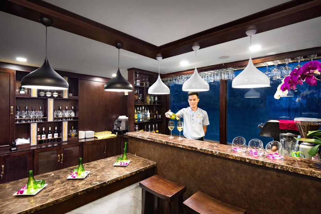 Elegant hotel bar with bartender pouring wine, luxurious wooden furnishings, and vibrant orchids.