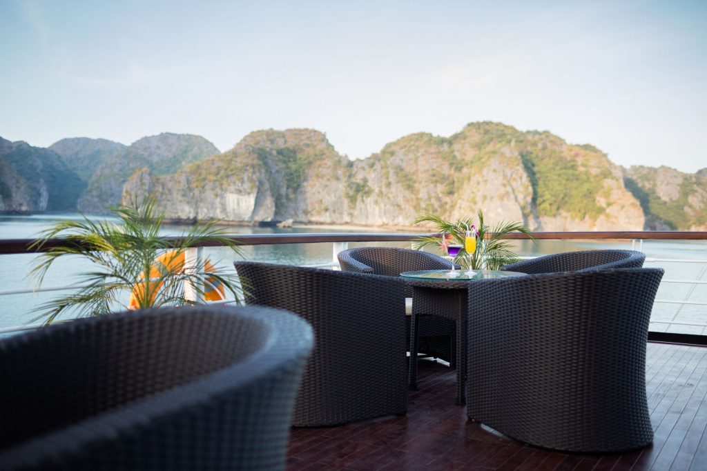 Panoramic view of a luxury cruise deck with wicker chairs and tropical plants, overlooking stunning limestone islands in Halong Bay.