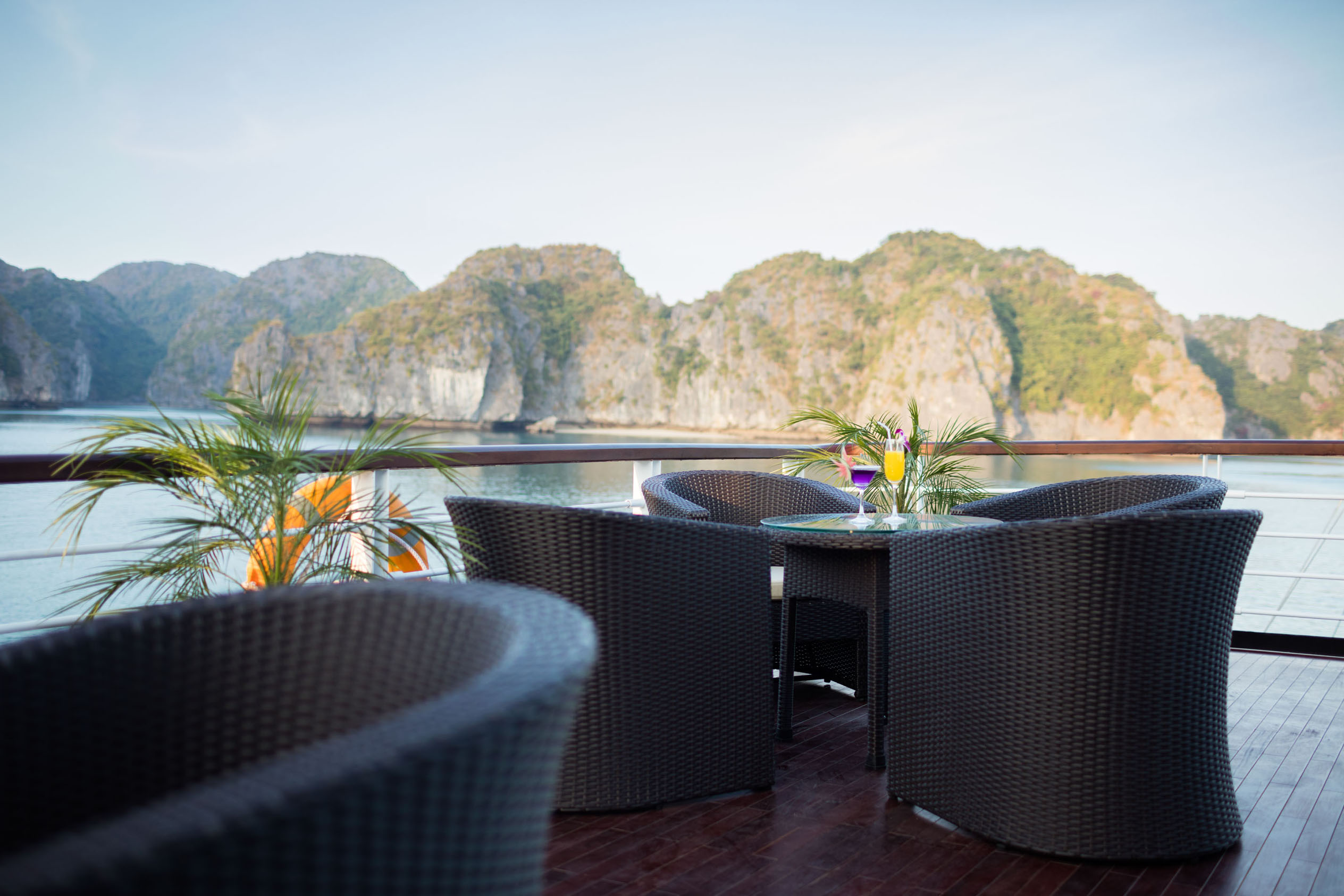 Panoramic view of a luxury cruise deck with wicker chairs and tropical plants, overlooking stunning limestone islands in Halong Bay.