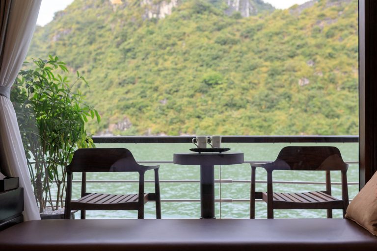 Serene lakeside view from a hotel room balcony with two cups on a table and lush green mountains in the background.