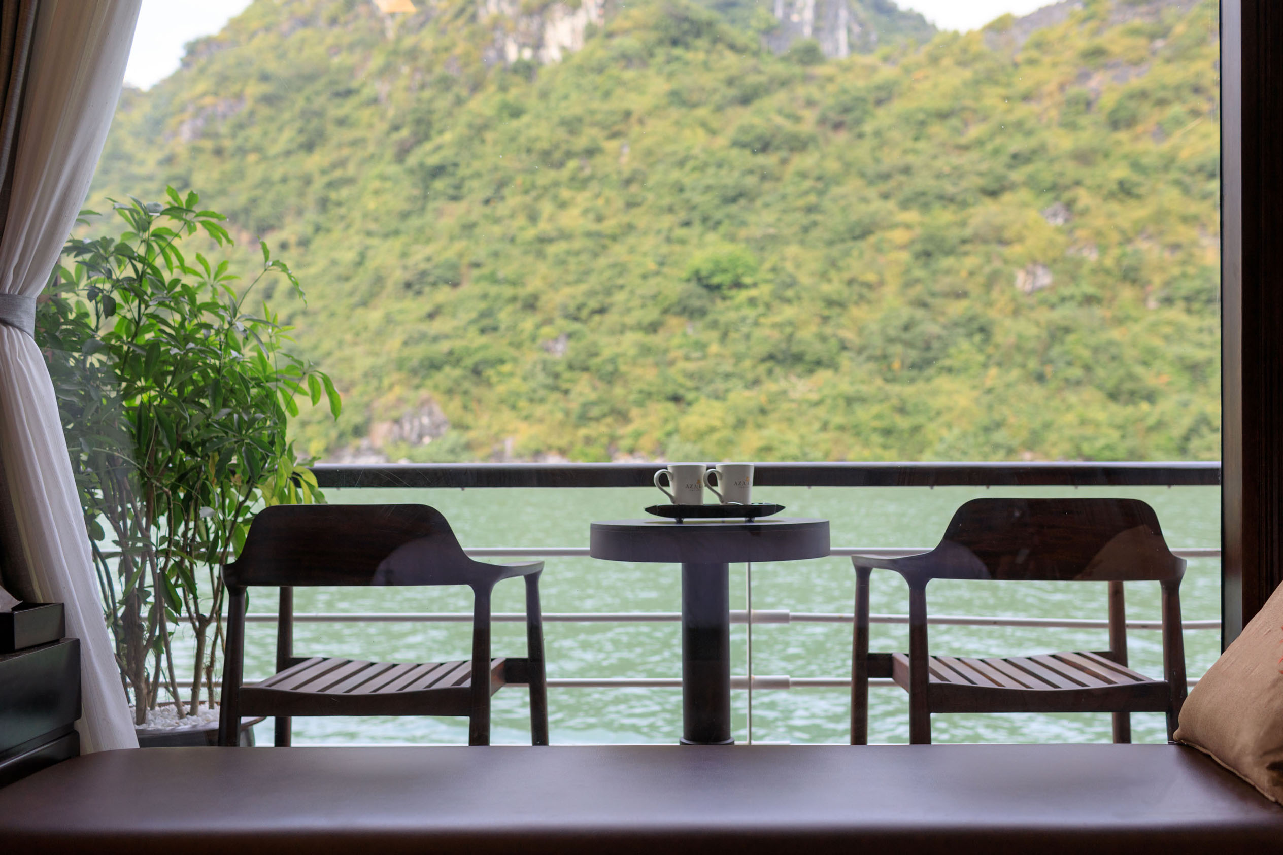 Serene lakeside view from a hotel room balcony with two cups on a table and lush green mountains in the background.