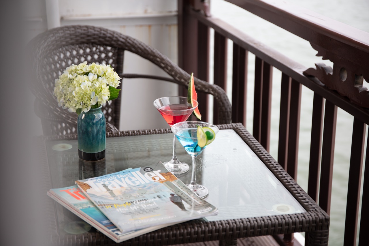 Elegant waterfront balcony with tropical cocktails, travel magazines, and a flower arrangement for a relaxing vacation vibe.