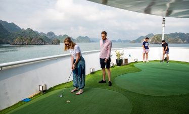 Gentlemen and women enjoying a game of mini-golf on the deck of a luxury cruise ship with scenic islands in the background.