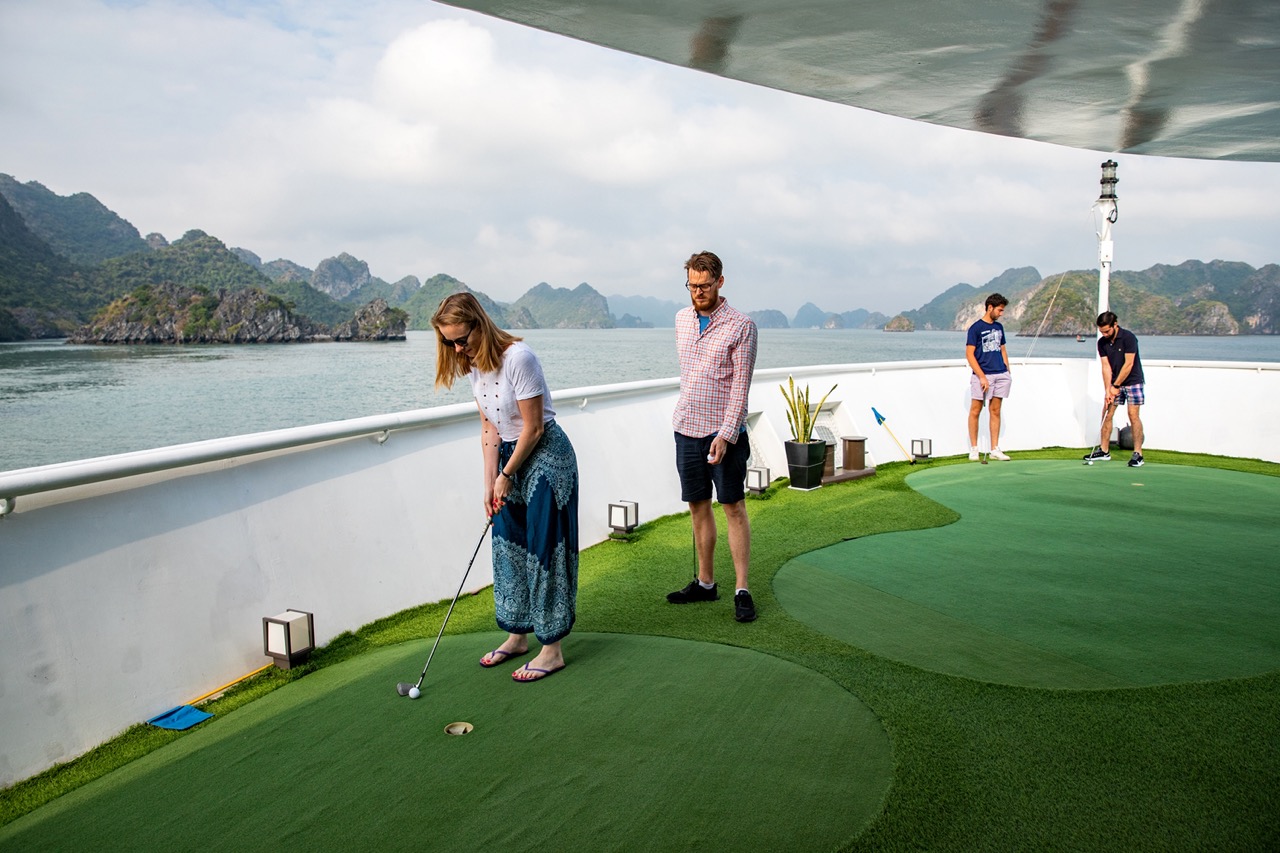 Gentlemen and women enjoying a game of mini-golf on the deck of a luxury cruise ship with scenic islands in the background.
