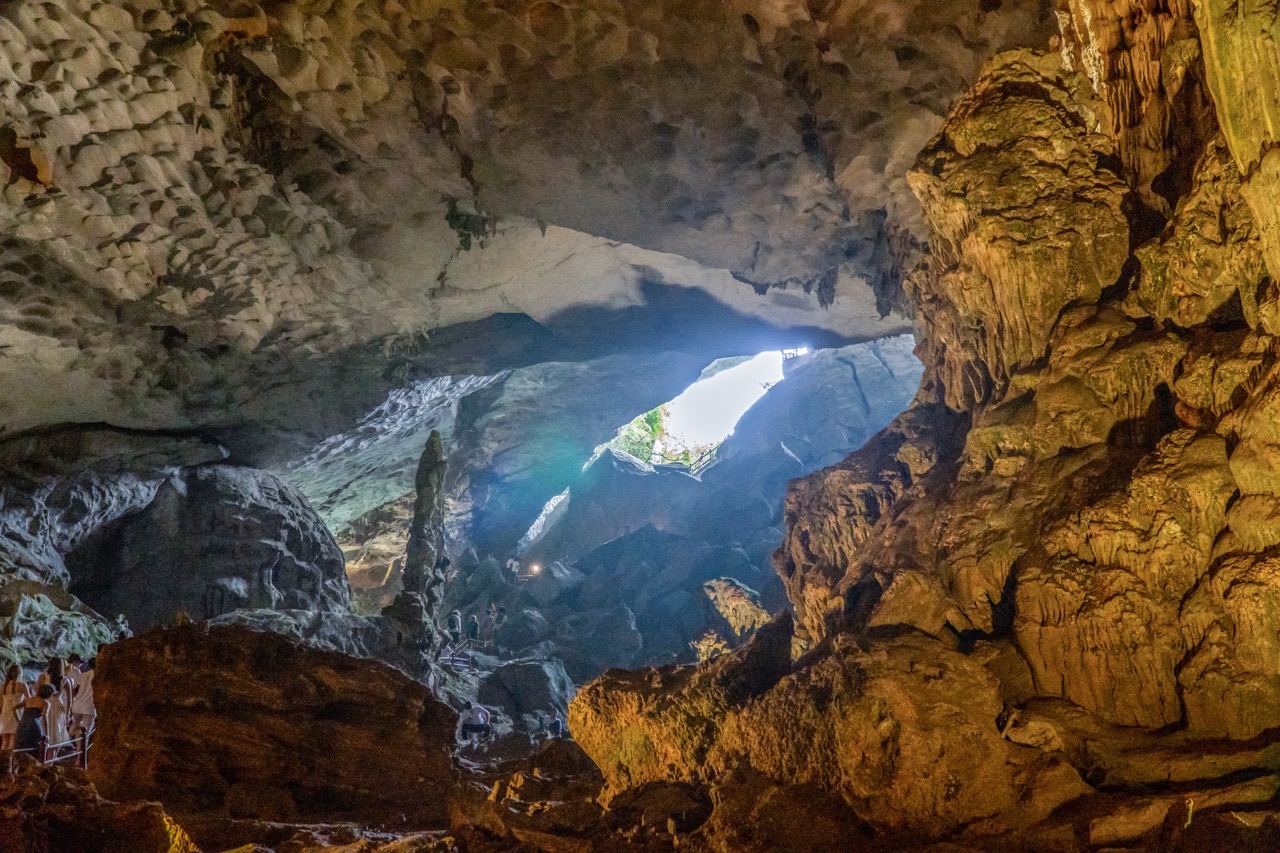 Imposing cave with sunlight rays, limestone formations, and tourists exploring inside.