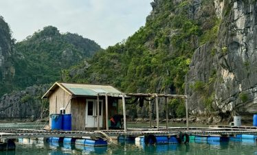 Floating fishing house on scenic water surrounded by limestone cliffs and lush greenery.