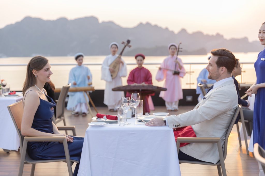 Romantic scene of a couple dining on Calista Cruise in Halong Bay during sunset, with traditional mu.