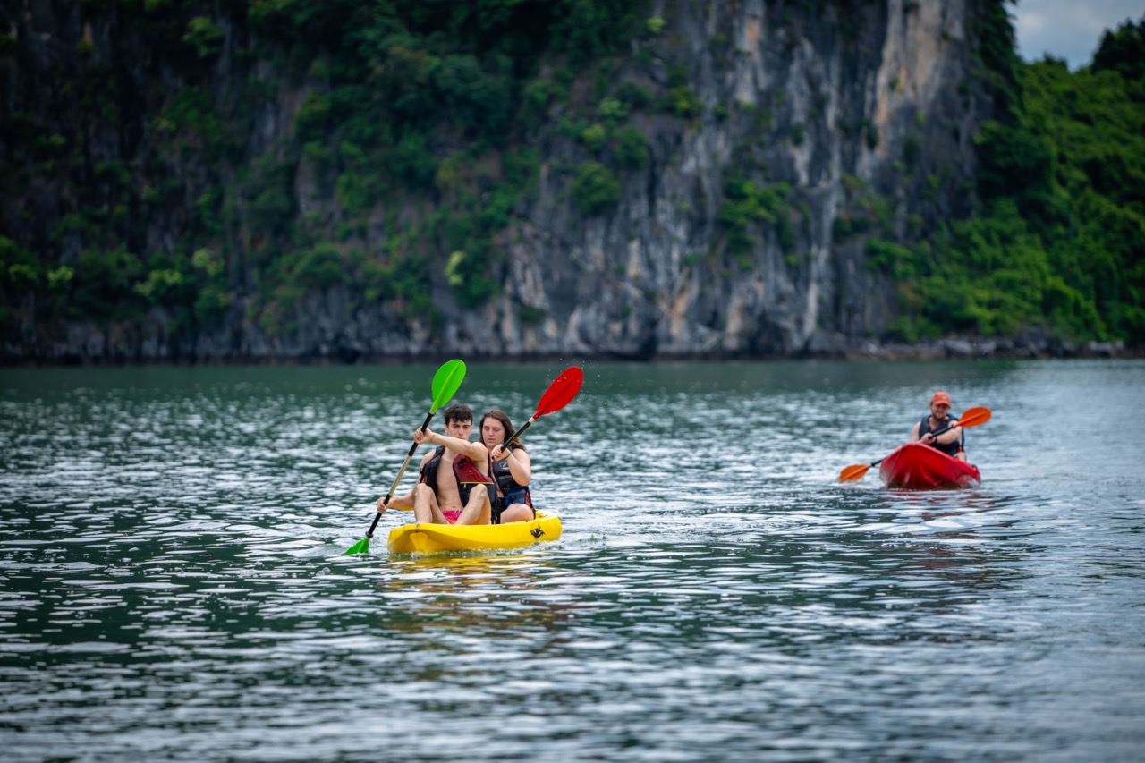 Kayakers paddling on calm Halong Bay waters with lush green cliffs in the background, enjoying a pea.
