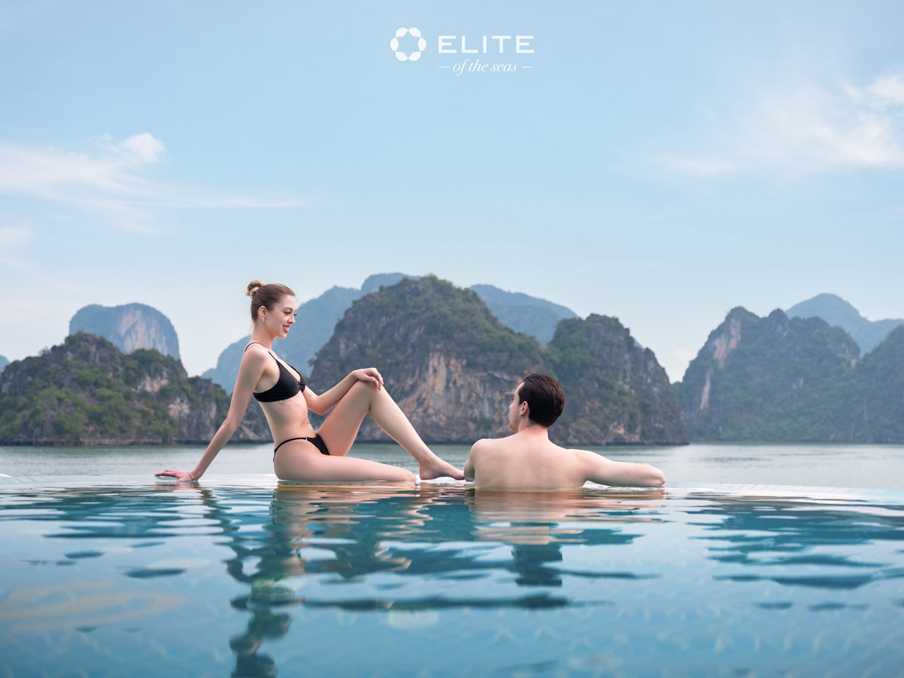 Relaxing couple on cruise balcony with stunning Halong Bay scenery, clear blue sky and limestone kar.