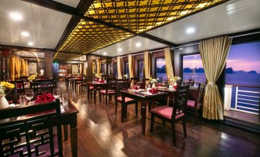 Luxurious interior of a cruise ship dining area with large windows overlooking Halong Bay at sunset.