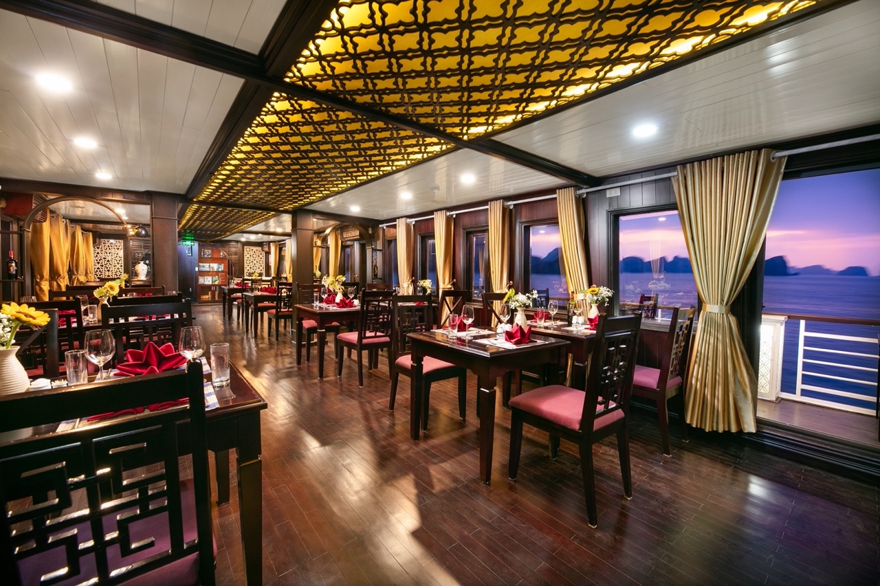 Luxurious interior of a cruise ship dining area with large windows overlooking Halong Bay at sunset.