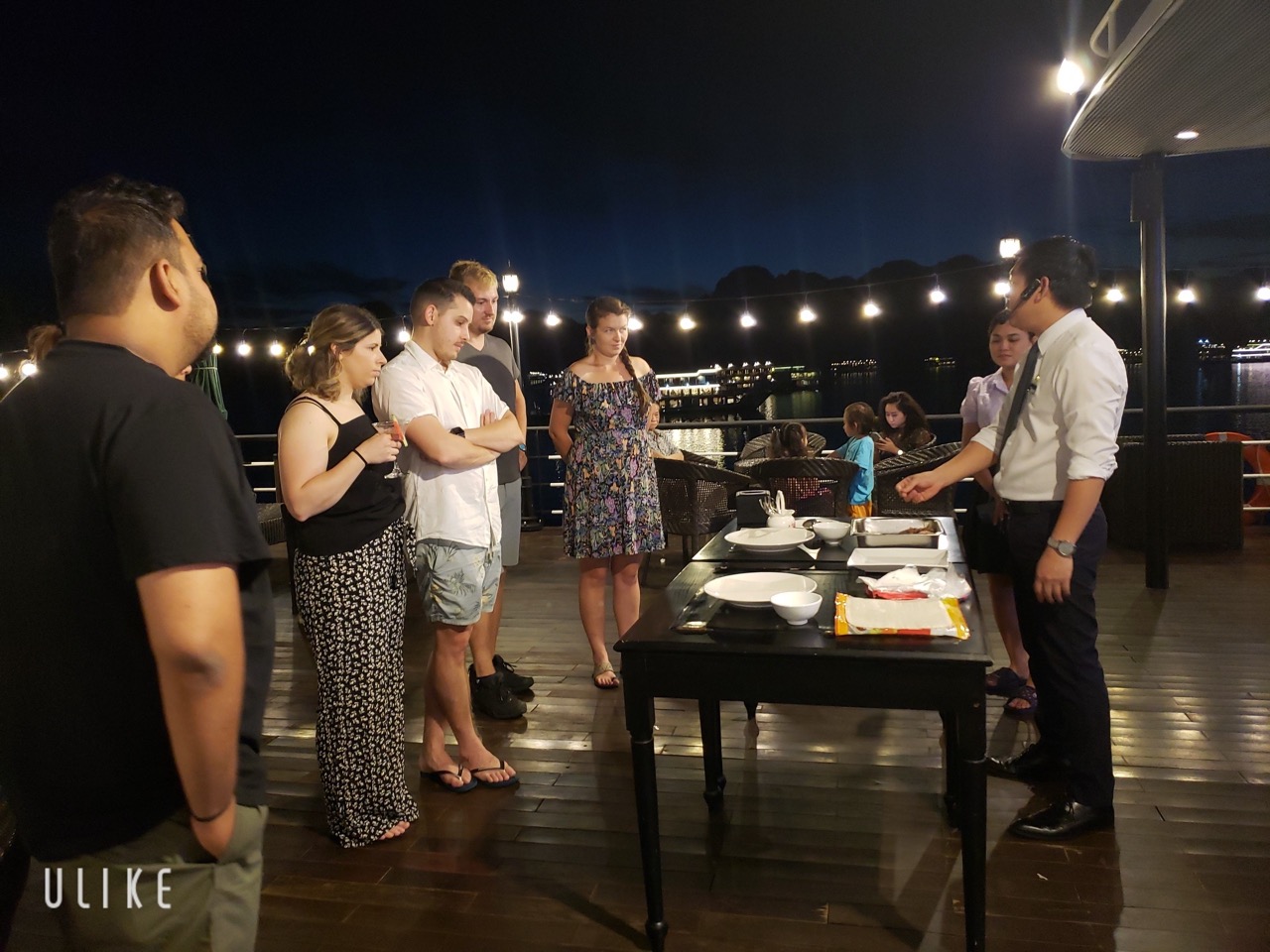 Halong Bay cruise dinner scene with guests enjoying a meal on the deck at night, scenic water views,.
