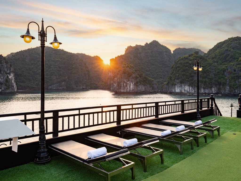 Sunset over Halong Bay with cruise balcony and lounge chairs, scenic mountain backdrop, peaceful eve.