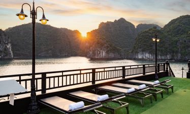 Sunset over Halong Bay with cruise balcony and lounge chairs, scenic mountain backdrop, peaceful eve.