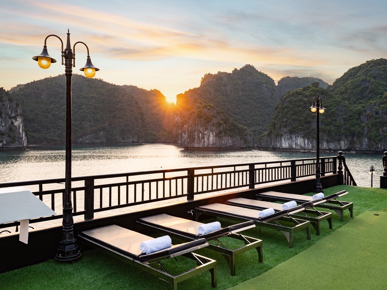Sunset over Halong Bay with cruise balcony and lounge chairs, scenic mountain backdrop, peaceful eve.