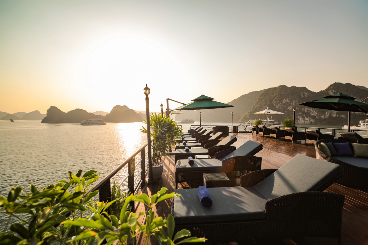Stunning sunset view from a luxury cruise balcony in Halong Bay, with lounge chairs and scenic limes.