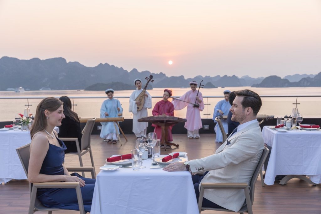 Romantic couple dining on a luxury cruise balcony in Halong Bay at sunset.