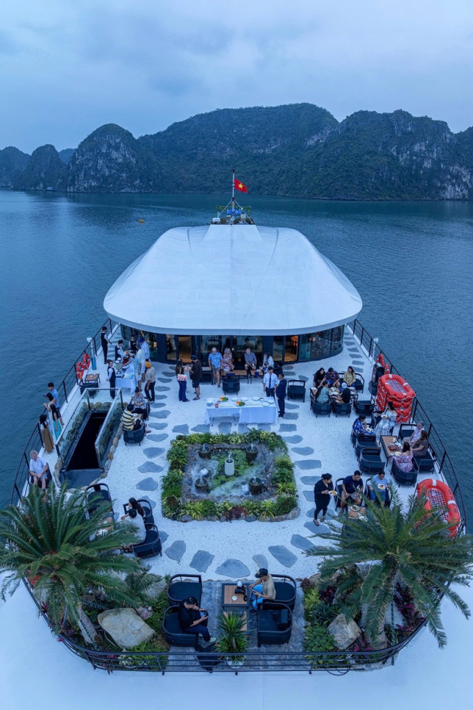 Aerial view of a restaurant boat with groups of people dining on the deck, surrounded by water and rocky islands; a small garden centerpiece is in the middle.