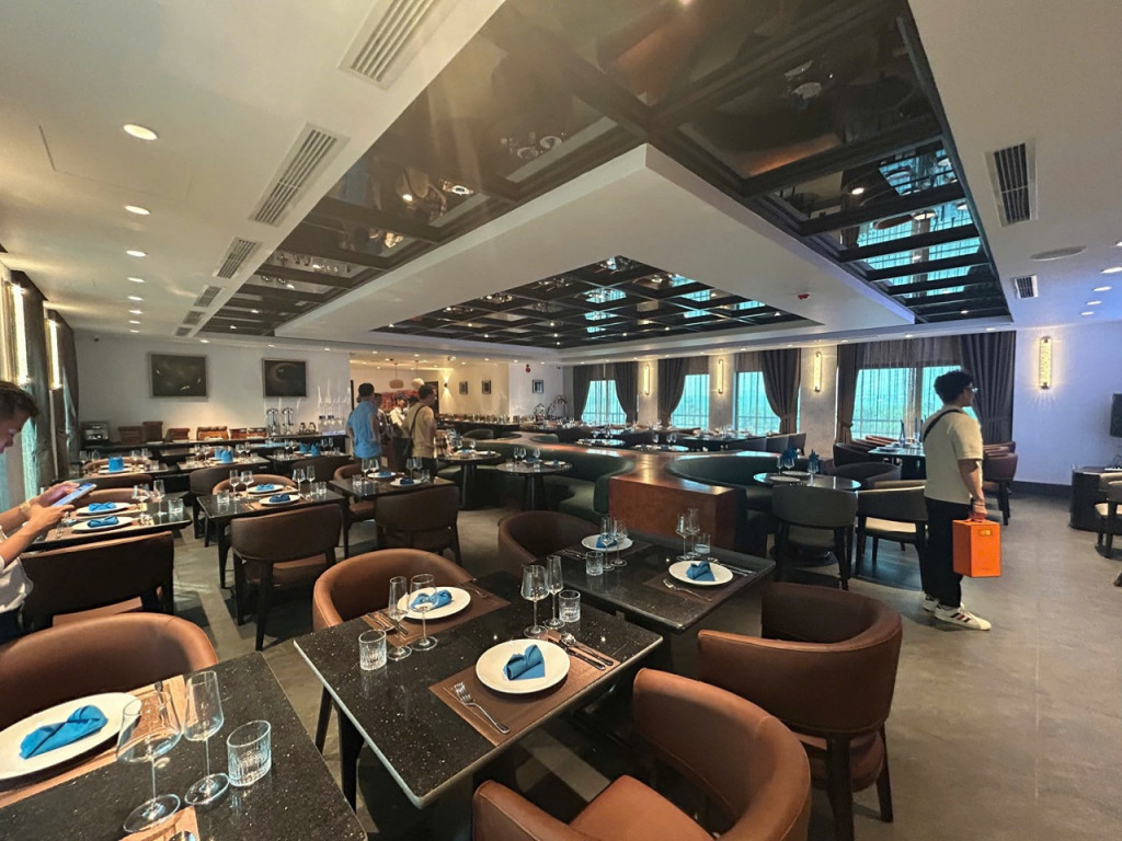 Upscale restaurant dining room with scattered round and square tables set with plates, glasses, and blue napkins; brown chairs and soft lighting.