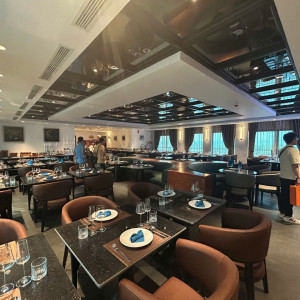 Upscale restaurant dining room with scattered round and square tables set with plates, glasses, and blue napkins; brown chairs and soft lighting.