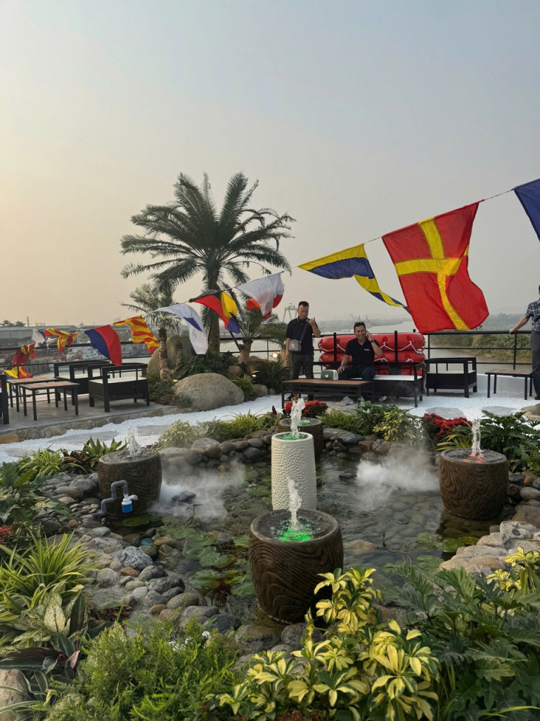 Rooftop garden with decorative fountains, rocks, and lush plants, set against colorful pennant flags and a seating area in the background.
