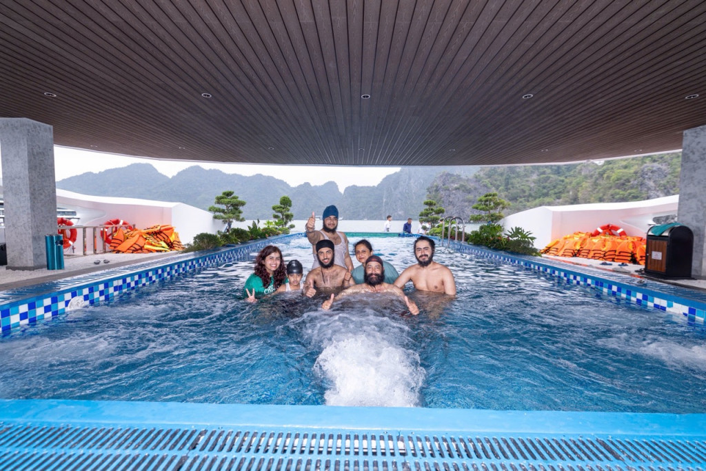 Group of seven people standing in a rectangular outdoor pool under a wooden covered deck, giving thumbs up with a mountainous landscape in the background.