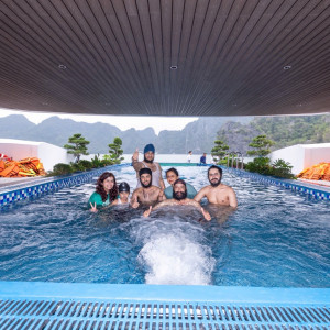 Group of seven people standing in a rectangular outdoor pool under a wooden covered deck, giving thumbs up with a mountainous landscape in the background.