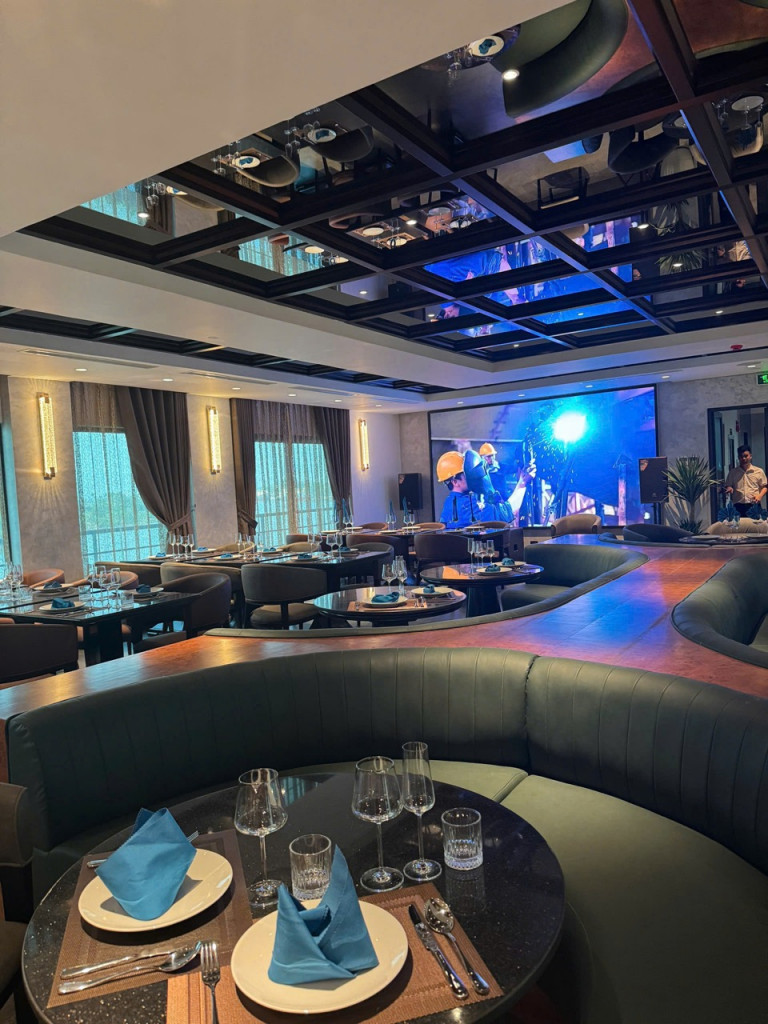 Upscale banquet room with curved leather booths, round tables set with blue napkins and glassware, and a large screen at the front for presentations/entertainment.