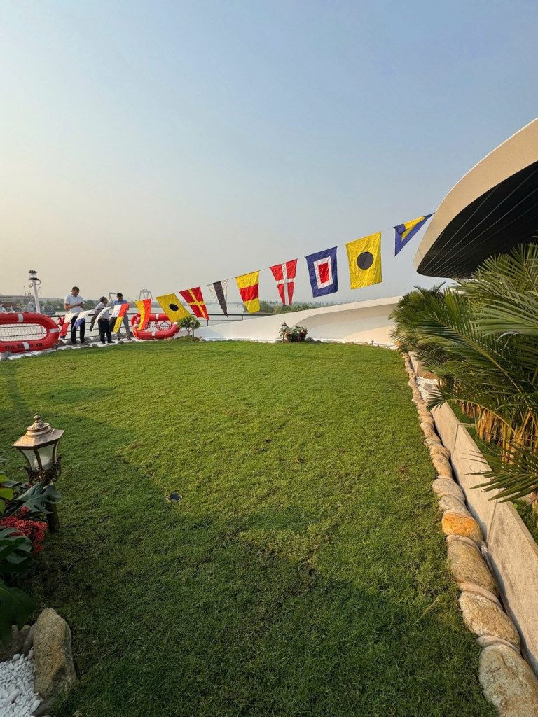 People stand along a line of colorful signal flags strung above a grassy terrace, with a curved white building and tropical plants on the right and water in the distance.