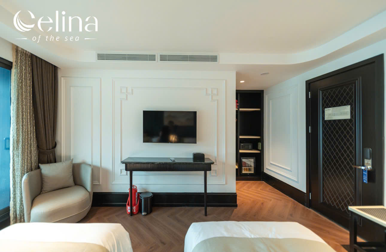 Hotel room with wall-mounted TV above a black console, beige seating chair, and two beds at the foreground; dark door on the right and built-in shelves near the entry. Logo 'Celina of the sea' at top left.