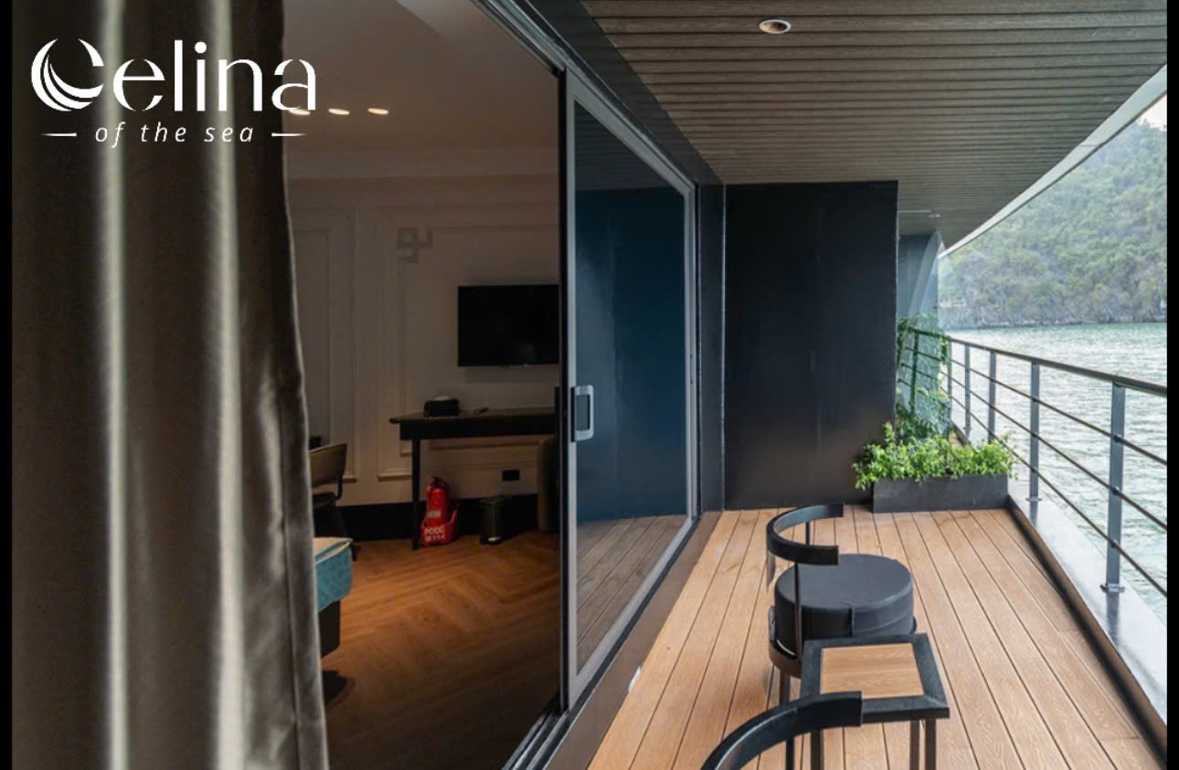 Balcony of a waterfront hotel with a wooden deck, glass sliding doors, and two black chairs, overlooking the water; logo 'celina of the sea' in the upper left.