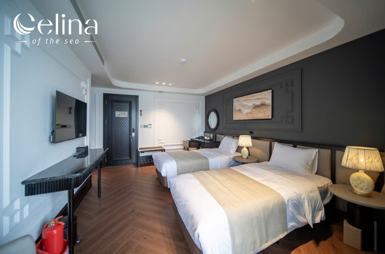 Hotel room with two tidy beds, beige bed runners, and warm lamps on round side tables; dark accent wall and framed art above the beds.