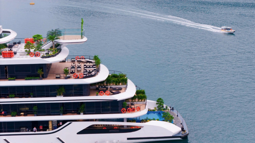 Multi-deck luxury yacht with terrace lounges, plants, and orange life rings, on calm blue water; a speedboat speeds by in the distance.