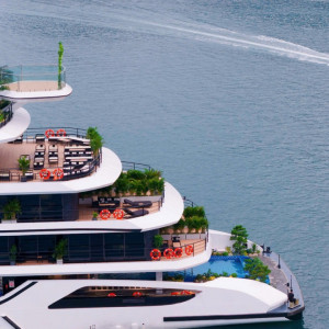 Multi-deck luxury yacht with terrace lounges, plants, and orange life rings, on calm blue water; a speedboat speeds by in the distance.