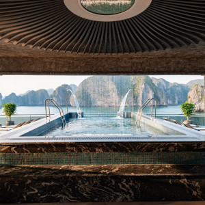 Luxurious indoor-outdoor pool framed by dark marble, with water jets and a bay of limestone karst rocks in the distance.