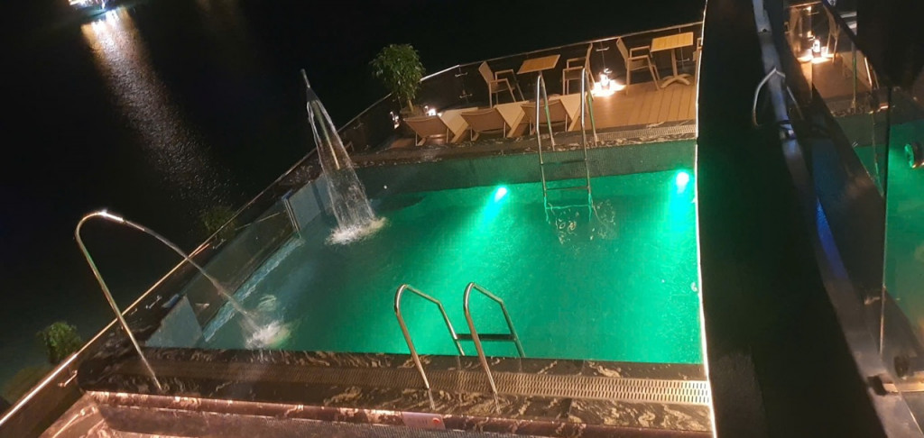 Nighttime pool with green lights, a fountain jet, and metal ladders; lounge chairs on the deck in the background.